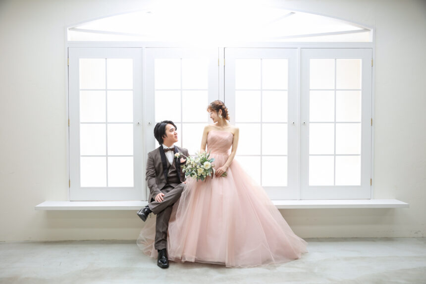 大きな窓辺で並ぶ新郎新婦のフォトウェディング・ピンクドレスが柔らかな光に映えるエレガントな一枚