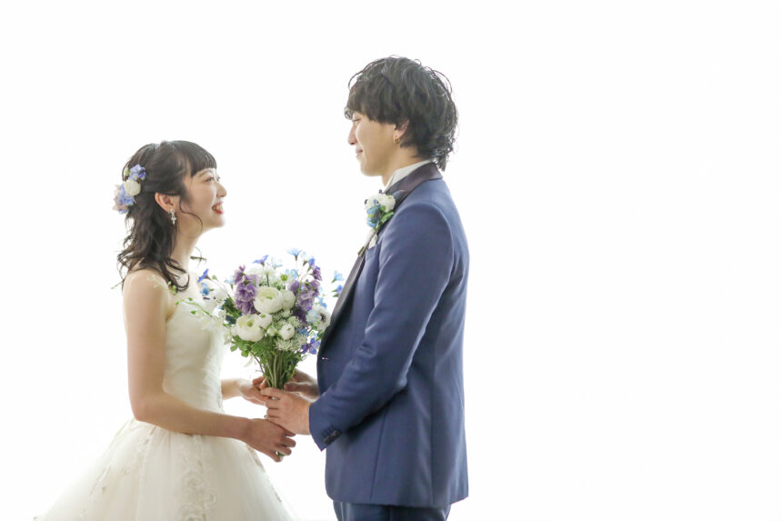 白い背景で向かい合う新郎新婦のフォトウェディング・白ドレスとブルータキシードが爽やかに映えるシンプルなスタジオショット