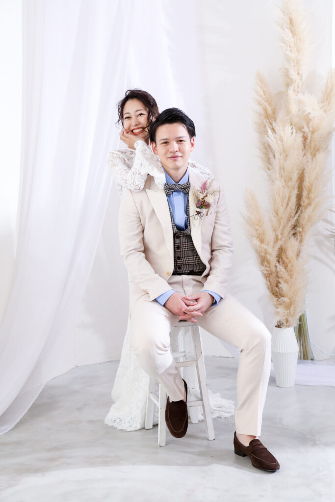 白いスタジオで椅子に座る新郎とその肩に手を添える新婦のフォトウェディング・柔らかな装飾と白ドレスが映える優しい雰囲気の一枚