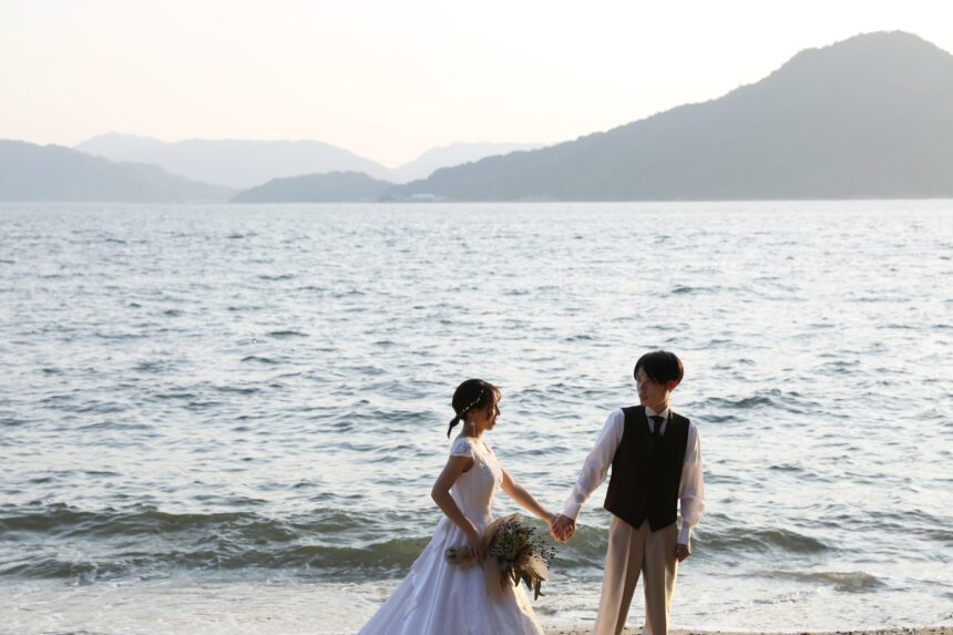 海辺の砂浜で手を取り合う新郎新婦のフォトウェディング・穏やかな海と遠くの山並みが優しく映るロマンチックな一枚