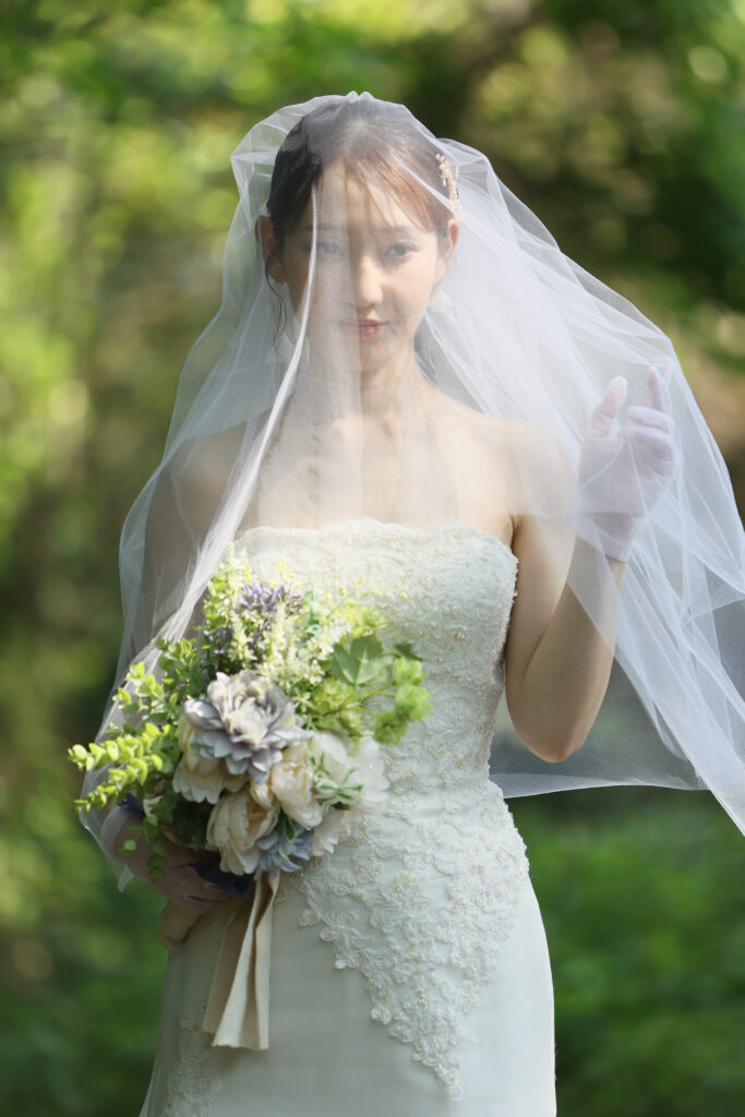 ベールをまといウェディングドレスでブーケを持つ花嫁のフォトウェディング・緑に囲まれた屋外で撮影した柔らかい雰囲気のショット