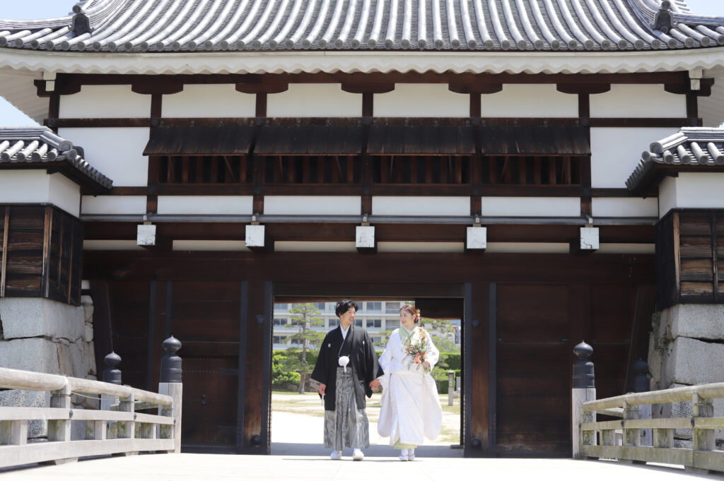 城門前の木橋で並んで歩く新郎新婦の和装フォト・黒紋付と白無垢が歴史的な建築に映える落ち着いた一枚