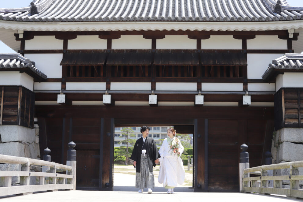 城門前の木橋で並んで歩く新郎新婦の和装フォト・黒紋付と白無垢が歴史的な建築に映える落ち着いた一枚