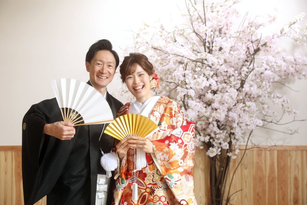 桜の装飾を背景に並ぶ和装の新郎新婦のフォトウェディング・華やかな和の雰囲気が広がる一枚