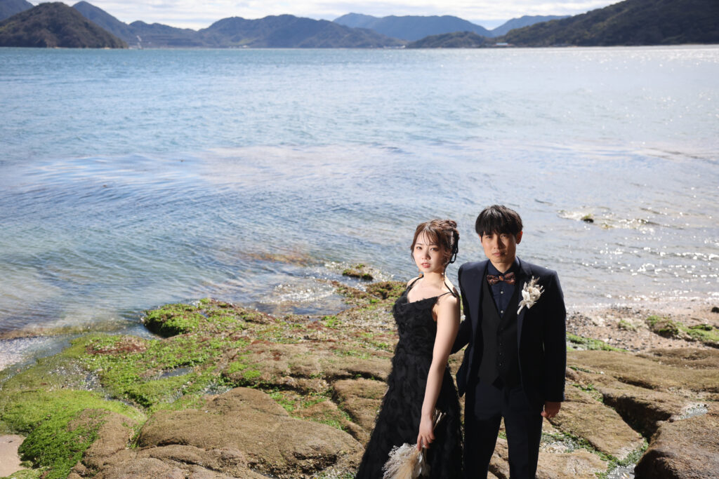 海辺の岩場で並ぶ新郎新婦のフォトウェディング・自然の景観が映えるシックなショット