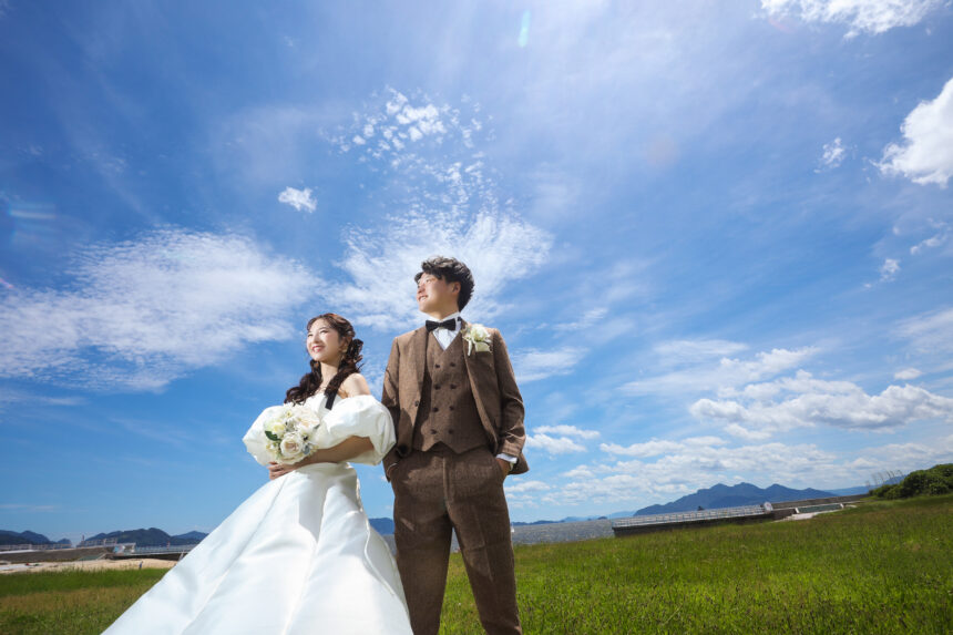 広い草原で並ぶ新郎新婦のフォトウェディング・白ドレスとブラウンタキシードが青空の下で映えるナチュラルなロケーションフォト