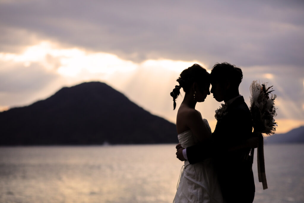 夕景の海辺で寄り添う洋装の新郎新婦のフォトウェディング・光に包まれた印象的なシルエットショット