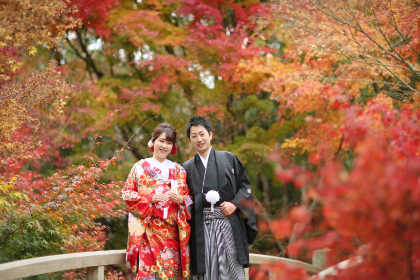 紅葉に包まれた橋の上で並ぶ新郎新婦の和装フォト・色打掛と黒紋付が秋の景色に映えるロケーションショット