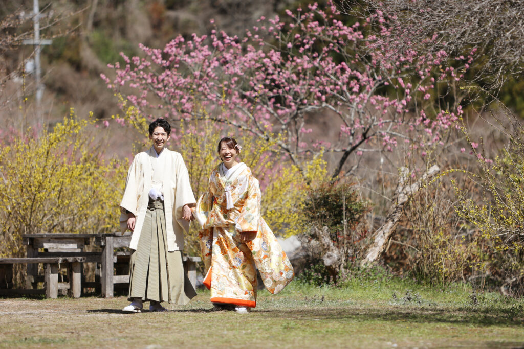 桃の花の下で並ぶ和装の新郎新婦のフォトウェディング・春の彩りが映える華やかな和装ショット