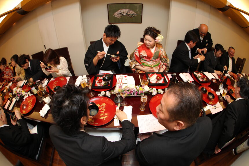 和装の新郎新婦を囲み、親族が会食を楽しむ披露宴の食事シーン。赤い漆器と花飾りが並ぶ落ち着いた和のテーブルコーディネート。