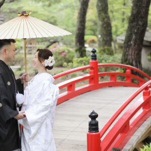 庭園の赤い太鼓橋の上で、黒紋付の新郎と白無垢の新婦が和傘を持って向き合う和装前撮り写真。緑豊かな景色が二人を包む。