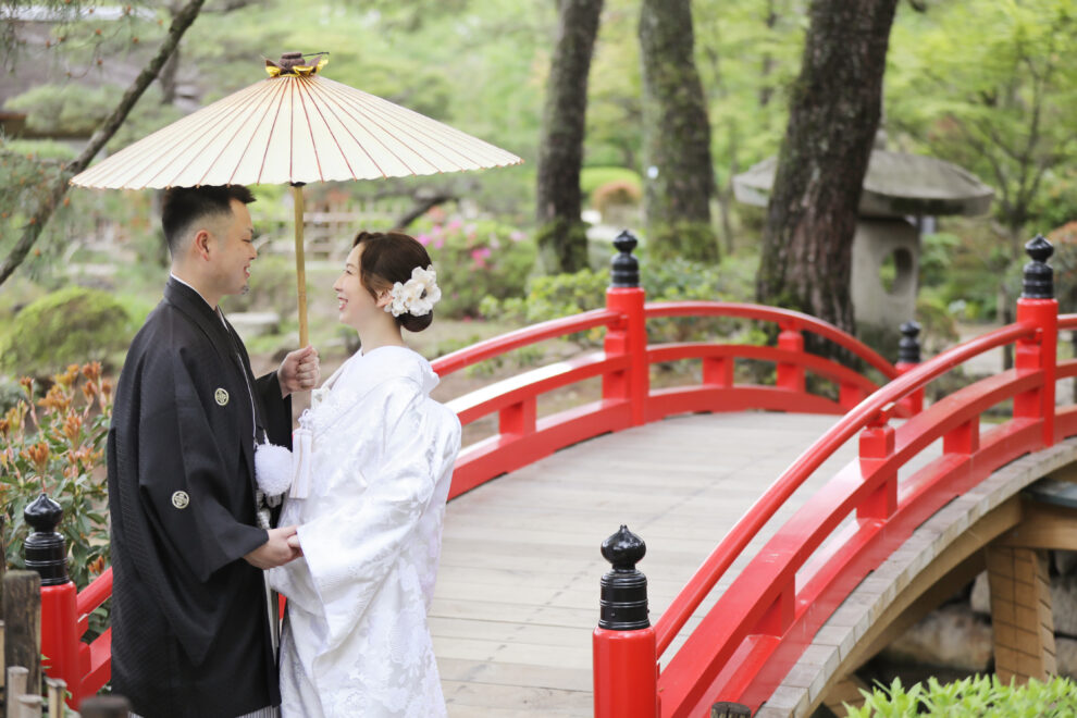 庭園の赤い太鼓橋の上で、黒紋付の新郎と白無垢の新婦が和傘を持って向き合う和装前撮り写真。緑豊かな景色が二人を包む。