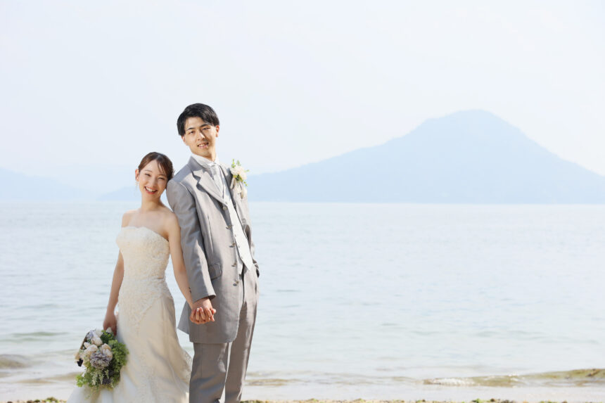 海辺で手を取り合う新郎新婦のフォトウェディング・白ドレスとグレータキシードが穏やかな海景色に映える爽やかなロケーションショット