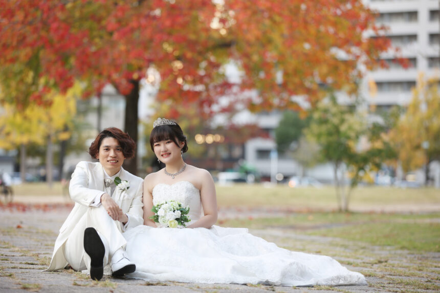 紅葉の公園で並んで座る洋装の新郎新婦の前撮り・秋の色彩に包まれた自然体ショット