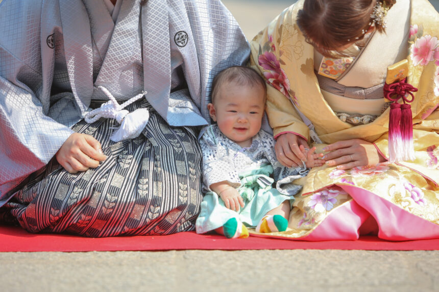 和装の新郎新婦に挟まれて座る赤ちゃんの記念フォト・赤い毛氈の上で家族の温もりを感じる一枚