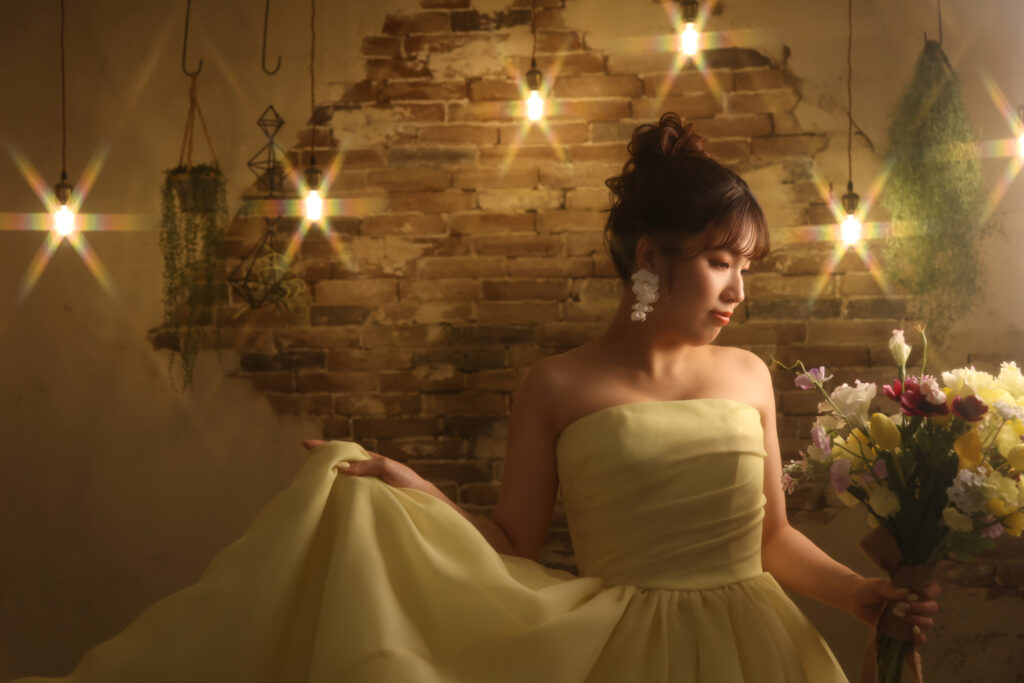レンガ壁と電球の柔らかな灯りに包まれた黄色ドレスの女性のフォトウェディング・カラフルなブーケが映えるロマンチックな一枚