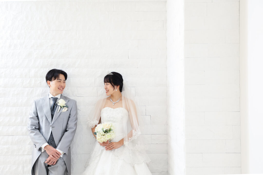 白いレンガ壁の前で向き合う新郎新婦のフォトウェディング・白ドレスとライトグレーのタキシードが映えるシンプルで上品な一枚