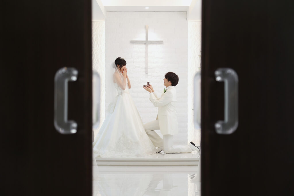 チャペルの扉越しにプロポーズする新郎新婦のフォトウェディング・白い空間に指輪ケースが映える印象的なシーン