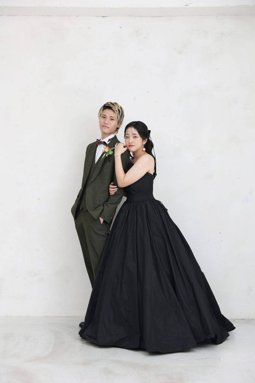 白壁の前で並ぶ新郎新婦のフォトウェディング・黒ドレスとグリーンスーツが映えるシックで洗練された一枚