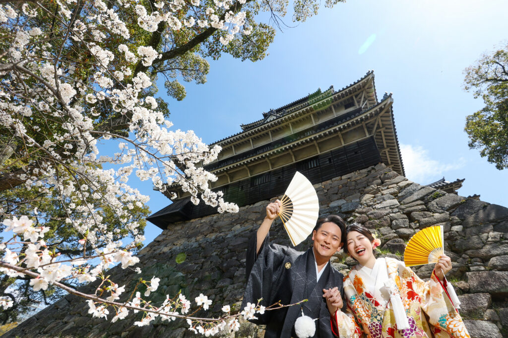 広島城の桜を背景に扇子を持つ和装の新郎新婦のフォトウェディング・春の城ロケが映える一枚