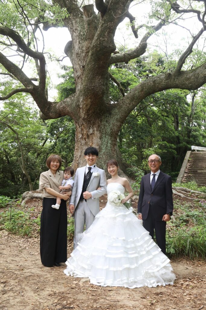 大きなクスノキを背景に、タキシード姿の新郎とウェディングドレス姿の新婦がご家族と並ぶ記念写真。自然豊かなロケーションが開放的な雰囲気を添える。