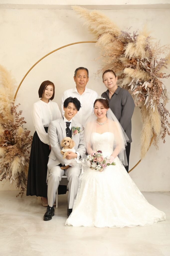 ドライフラワーで彩られたフォトスタジオで、タキシード姿の新郎とウェディングドレス姿の新婦が愛犬を抱き、ご家族と並ぶ記念写真。ナチュラルな装飾が温かな雰囲気を演出する。
