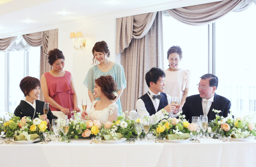 会食会場で、タキシード姿の新郎新婦が両親や家族と歓談を楽しむ会食付きフォトウェディングの様子。華やかな装花が並ぶテーブルが祝福の時間を彩る。
