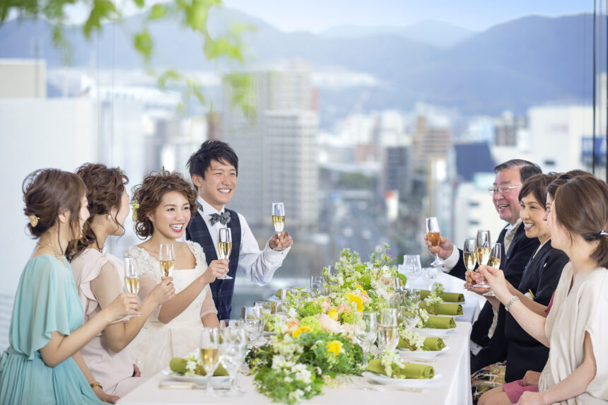 レストランの会場で、タキシード姿の新郎とウェディングドレス姿の新婦が家族と乾杯を楽しむ会食付きフォトウェディングの様子。窓の外に広がる街並みが開放的な雰囲気を演出する。